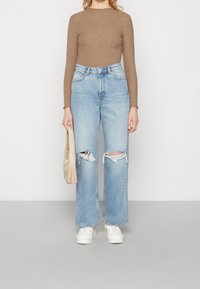 Maglietta marrone a coste a maniche lunghe, jeans a gamba larga blu chiaro con ampi strappi, sneakers bianche e una piccola borsa beige.