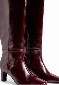 Burgundijas ceļgaliem augstas ādas zābaki ar spīdīgu apdari, smailu pirkstu un 3 collu blokveida papēdi. Aprīkoti ar sānu rāvējslēdzēju vieglai uzvilkšanai.