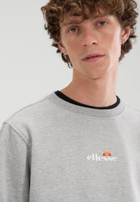 Sudadera gris con cuello redondo, que presenta un logo colorido "ellesse" en el pecho, fabricada con algodón suave y acabado texturizado.