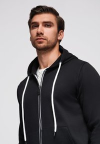 Felpa nera con zip, cappuccio con coulisse e accenti bianchi, caratterizzata da una texture liscia e tasche frontali. Il modello indossa una T-shirt grafica sotto.