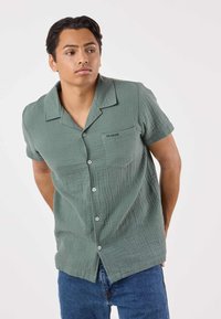 Camicia a maniche corte con colletto in tessuto verde chiaro strutturato, con taschino frontale con il logo "SEASIDE" e bottoni bianchi. Abbinata a jeans blu.