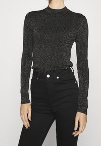 Czarna prążkowana bluzka z długim rękawem wykonana z błyszczącego materiału, z wysokim kołnierzem i dopasowanym krojem, zestawiona z czarnymi, wysokim stanem jeansami.