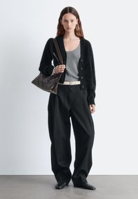 Schwarzer flauschiger Cardigan, graues Tanktop, schwarze Hosen mit weitem Bein, weißer Gürtel und dunkle geflochtene Tasche. Glatte Textur mit Knopfverschluss und langen Ärmeln.