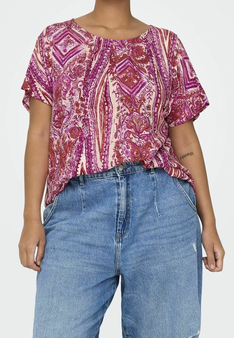Short-sleeve, pink blouse with a vibrant purple and red geometric floral pattern; relaxed fit; worn with light blue high-waisted jeans.