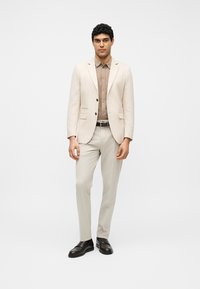 Άνδρας που φοράει μπεζ κοστούμι με καφέ πουκάμισο με κουμπιά και μαύρα παπούτσια, στέκεται μπροστά σε απλό λευκό φόντο.