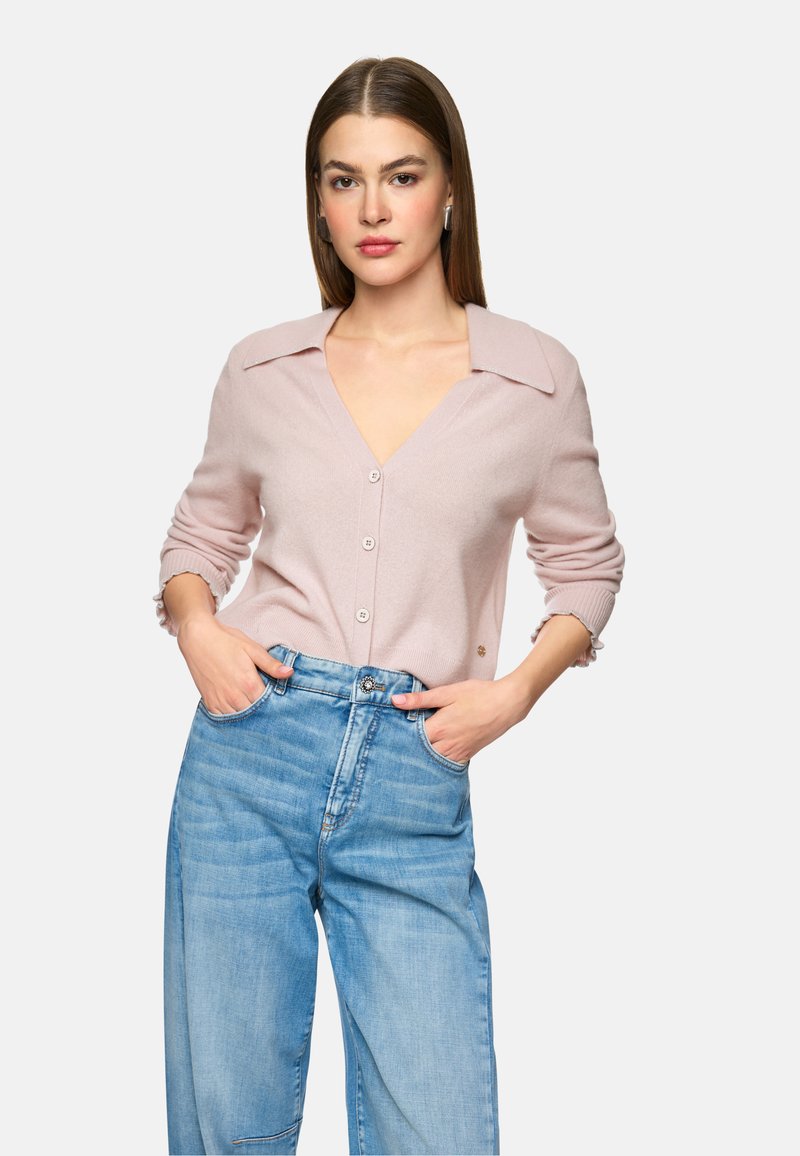 Helles rosa gestricktes Cardigan mit V-Ausschnitt und großem Kragen, mit Knopfverschlüssen und gerippten Ärmelakzenten. Getragen mit hellblauen Jeans im weit geschnittenen Stil.