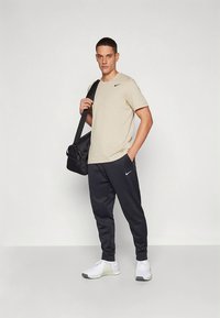 Camiseta beige de manga corta con el logo de Nike, pantalones joggers negros, zapatillas deportivas blancas y llevando una bolsa de gimnasio negra, de pie contra un fondo claro.