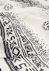 Tissu blanc avec des motifs géométriques et ponctués noirs, comprenant des lignes, des triangles et de petits dessins circulaires évoquant le soleil.