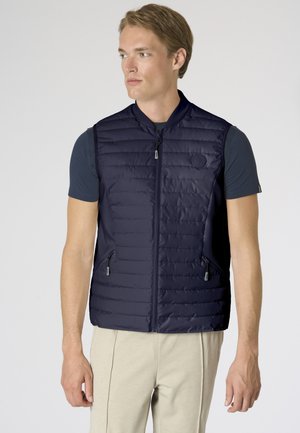 Uomo che indossa un gilet trapuntato blu navy con cerniera sopra una camicia scura a maniche corte e pantaloni beige, guarda verso sinistra su uno sfondo semplice.