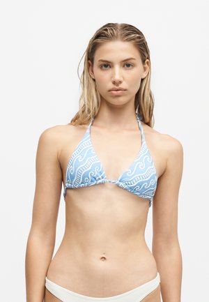 Jeune femme aux cheveux blonds mouillés portant un haut de bikini bleu à motif et un bas de bikini blanc, faisant face à l'avant sur un fond blanc.
