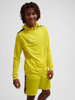 Junge Person lächelt, trägt einen leuchtend gelben Zip-Hoodie und passende Shorts mit schwarzen Chevron-Streifen an Schultern und Seiten.