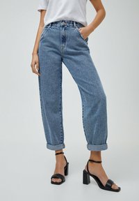 Jeans de tiro alto en azul claro con un ajuste relajado, presentan dobladillos enrollados y un diseño clásico de cinco bolsillos. Combinados con sandalias de tacón negras.