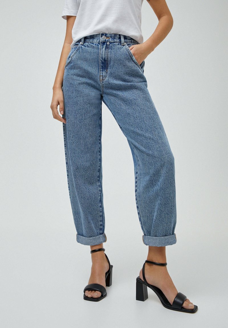 Jeans de tiro alto en azul claro con un ajuste relajado, presentan dobladillos enrollados y un diseño clásico de cinco bolsillos. Combinados con sandalias de tacón negras.