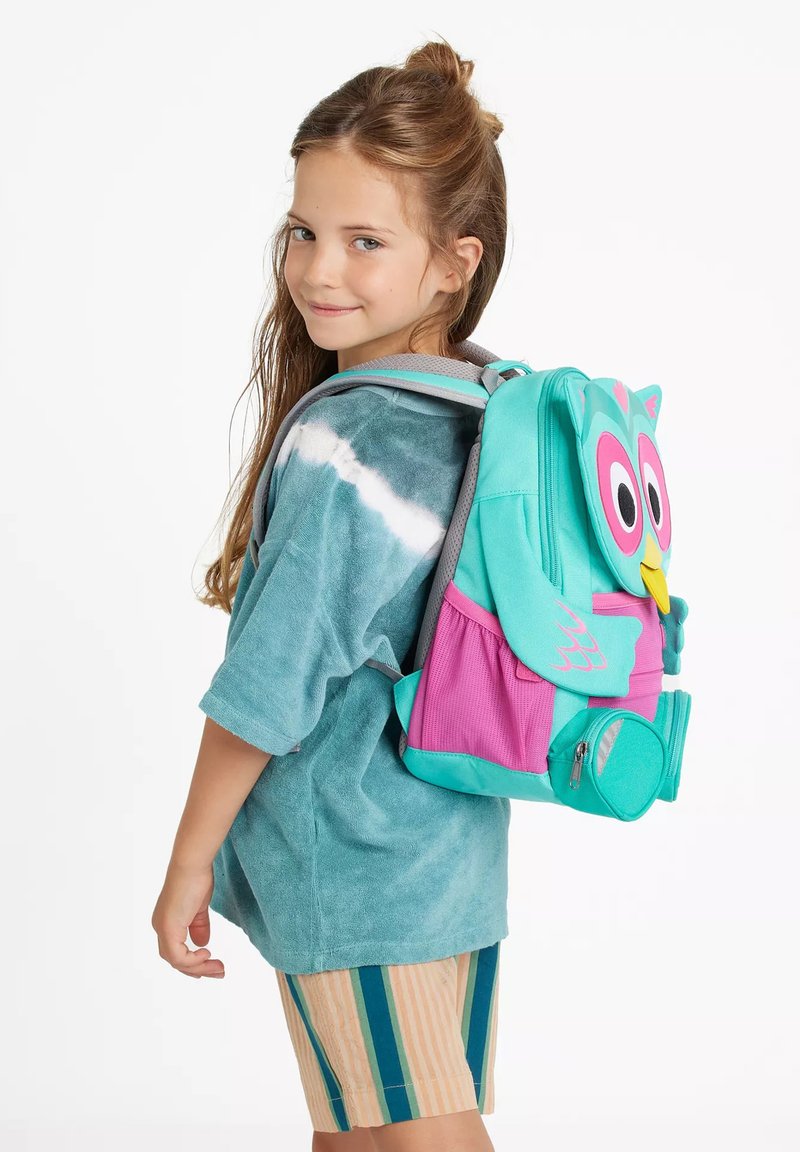 Junges Mädchen mit langen Haaren, trägt ein Türkisfarbendes Batik-Shirt und gestreifte Shorts, lächelt mit einem bunten, eulenförmigen Rucksack.