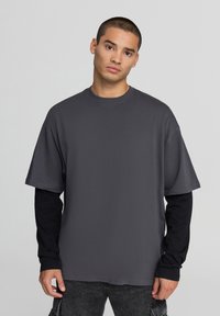T-shirt grigio oversize con maniche corte, realizzato in tessuto morbido, indossato sopra una maglietta nera a maniche lunghe. Presenta un collo rotondo.