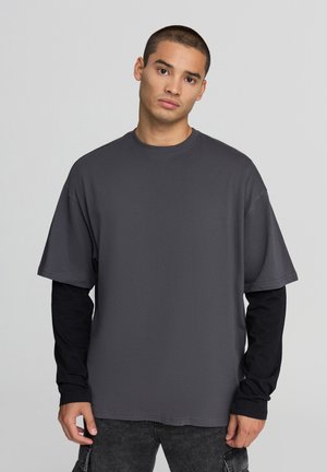 T-shirt grigio oversize con maniche corte, realizzato in tessuto morbido, indossato sopra una maglietta nera a maniche lunghe. Presenta un collo rotondo.