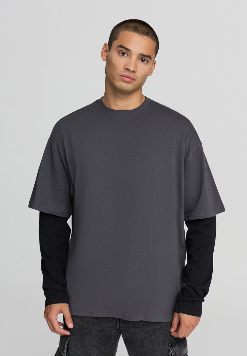 T-shirt grigio oversize con maniche corte, realizzato in tessuto morbido, indossato sopra una maglietta nera a maniche lunghe. Presenta un collo rotondo.
