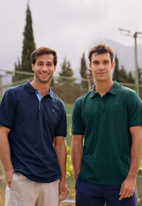 Kaksi miestä seisoo vierekkäin, pukeutuneina puuvillapolo-paitoihin; toinen tummansininen vaaleansinisellä kauluksella, toinen tummanvihreä, molemmissa on lyhyet hihat.