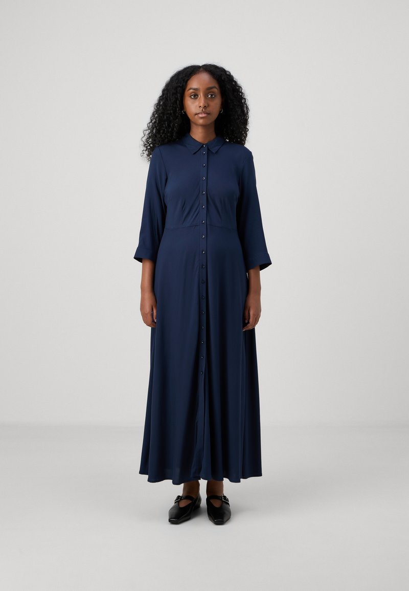 Jūras zila kleita ar garām piedurknēm, ar pogu aizdari, apkakli un plūstošu siluetu; izgatavota no gludas auduma un piedāvā brīvu piegriezumu.