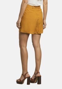 Pantalones cortos de lino mostaza con dobladillo doblado, bolsillos traseros y una cintura alta, combinados con sandalias marrones de tacón alto con tacón grueso.