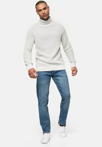 Witte gebreide trui met een hoge kraag, gecombineerd met blauwe jeans en witte sneakers. De trui heeft een gestructureerd patroon en geribde manchetten.