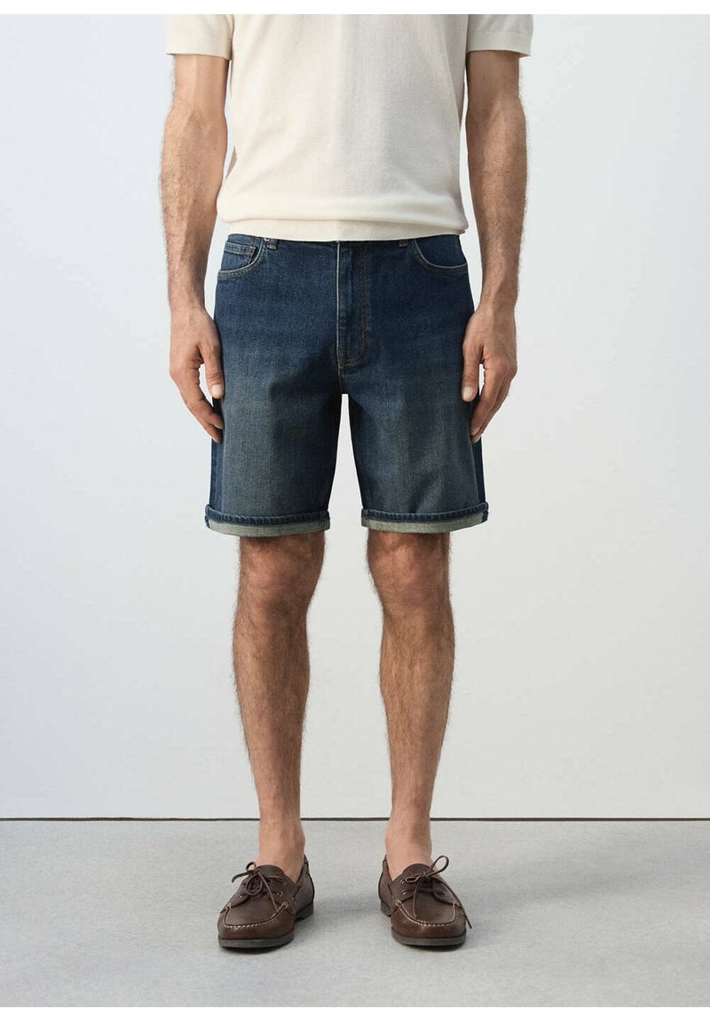 Homme portant un short en jean bleu foncé retroussé, une chemise courte crème et des chaussures en cuir marron à lacets, debout sur un sol gris.