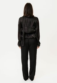 Melna satīna blouse ar puff piedurknēm, sasaistītas aizmugurē, un melnām platām biksēm. Gluda tekstūra ar spīdīgu apdari. Aizmugures skats.