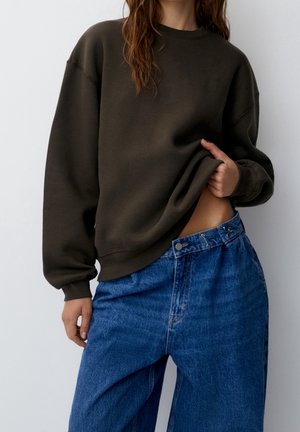 Femme soulevant légèrement un sweat-shirt marron foncé, montrant le ventre, portant un jean denim bleu ample avec taille haute et détails de boutons.