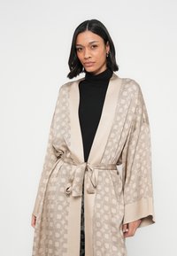 Kimono beige à motif avec une finition satinée, doté d'une ceinture à nouer à la taille et de manches larges, superposé à un col roulé noir.