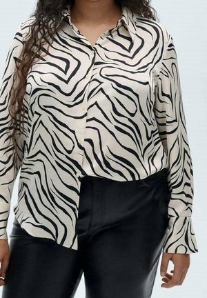 Kvinde iført en cremefarvet skjorte med knapper og sorte abstrakte zebrastriber samt sorte bukser med høj talje foran en ensartet baggrund.