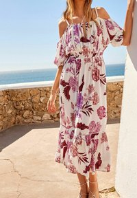 Donna che indossa un vestito bianco con spalle scoperte e un motivo floreale viola, in piedi accanto a un muro di pietra mentre guarda il mare in una giornata soleggiata.