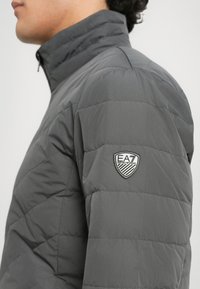 Sidevinkel af en person iført en grå quiltet jakke med EA7 Emporio Armani logo-patch på den øverste ærme.