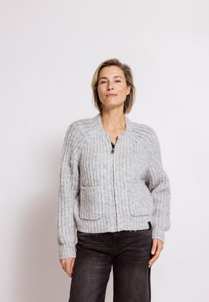 Woman standing wearing a light gray knitted zip-up sweater with pockets and dark jeans against a plain white background.