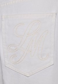 Bílá džínová kapsa s béžovými vyšitou skriptovou značkou „LM“ v dekorativním kurzívním stylu na látkovém pozadí.