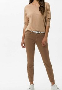 Schlanke, hellbraune Jeans mit zwei Fronttaschen, kombiniert mit einem beigen Langarmshirt und einem weißen Gürtel, abgerundet mit weißen Sneakers.
