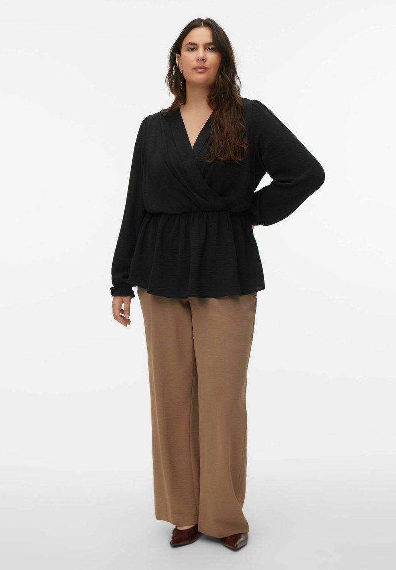 Blouse noire cache-cœur à manches longues avec taille élastique, associée à un pantalon beige à jambes larges et des ballerines marron pointues, sur fond blanc.