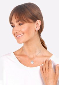 Zilveren ketting met ronde hangers, gedragen over een wit shirt. Het model heeft kort bruin haar en een subtiele oorbel.
