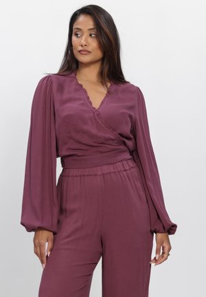 Woman wearing a long-sleeve, deep mauve wrap blouse and matching high-waisted pants, looking to her left against a plain background.