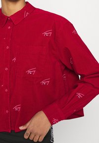Red corduroy shirt featuring a button-up design, single chest pocket, and repeated white logo accents throughout. Collared with long sleeves.