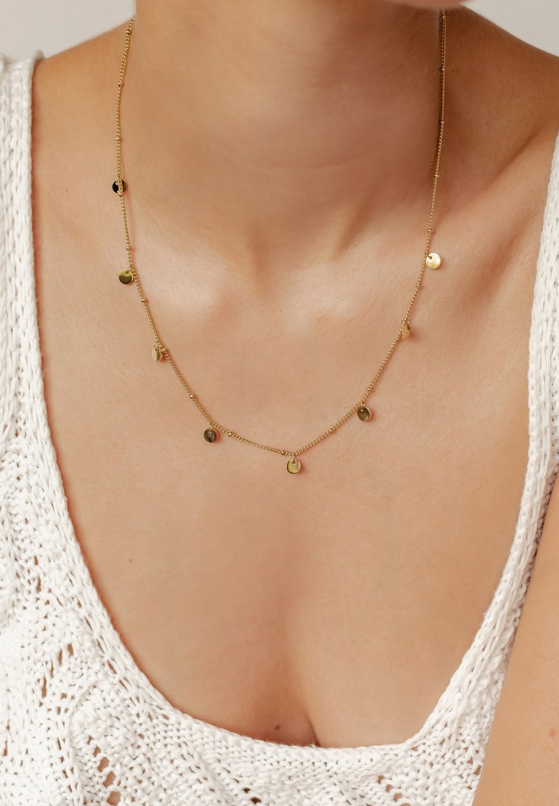 Gold chain necklace with small circular pendants, featuring a smooth texture and a delicate design, worn against a light, knit top.