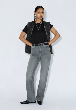 Grå vidde jeans, sort t-shirt og en sort denimvest med en sølvkæde halskæde. Modellen står med hænderne på hofterne mod en ensfarvet baggrund.