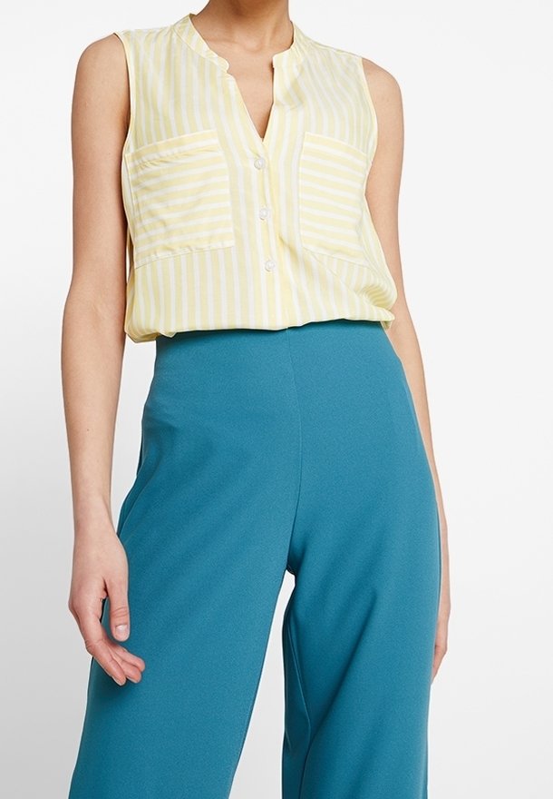 Femme portant un chemisier sans manches à rayures jaunes et blanches, boutonné, rentré dans un pantalon large taille haute bleu sarcelle.