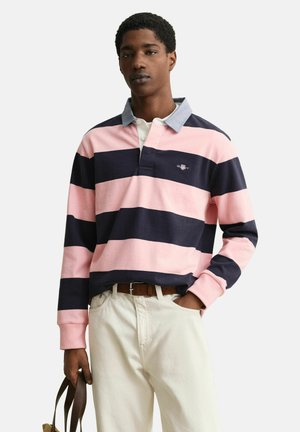 Mann trägt ein rosa und marineblau gestreiftes Rugbyhemd mit hellblauem Kragen, weiße Hose und hält einen braunen Leder-Taschenträger.