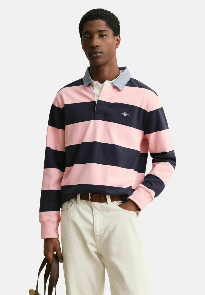 Uomo che indossa una maglia da rugby a righe rosa e blu navy con colletto azzurro, pantaloni bianchi e tiene una tracolla di borsa in pelle marrone.