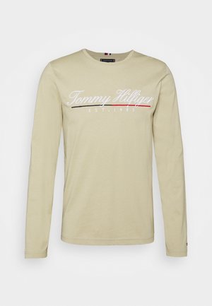 Beżowa koszula z długim rękawem z napisem "Tommy Hilfiger EST. 1985" w białym skrypcie oraz czarnym i czerwonym podkreśleniem na klatce piersiowej.