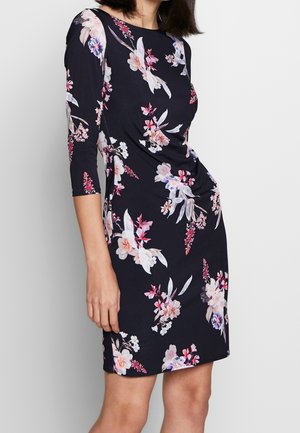 Femme portant une robe ajustée de couleur bleu marine avec des imprimés floraux rose, blanc et violet, à manches trois quarts, sur un fond uni.
