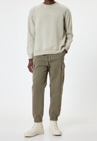 Mężczyzna w jasnobeżowej bluzie, oliwkowych spodniach cargo z kieszeniami z klapkami oraz białych sneakersach za kostkę, stoi z jedną ręką w kieszeni.