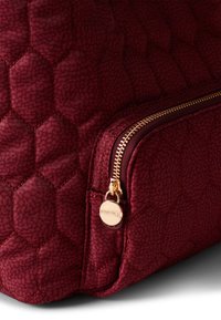 Deep red quilted bag featuring a hexagonal pattern, gold zipper hardware, and a round logo tag attached to the zip pull.