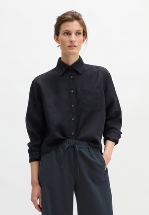 Vrouw met gebonden haar, gekleed in een zwart, geknoopt overhemd met lange mouwen en donker, loszittend broek met trekkoord, staand tegen een effen achtergrond.