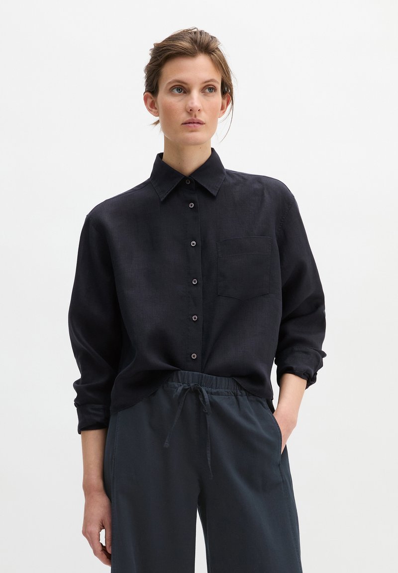 Donna con i capelli raccolti che indossa una camicia nera a maniche lunghe abbottonata e pantaloni scuri larghi con coulisse, in piedi davanti a uno sfondo semplice.